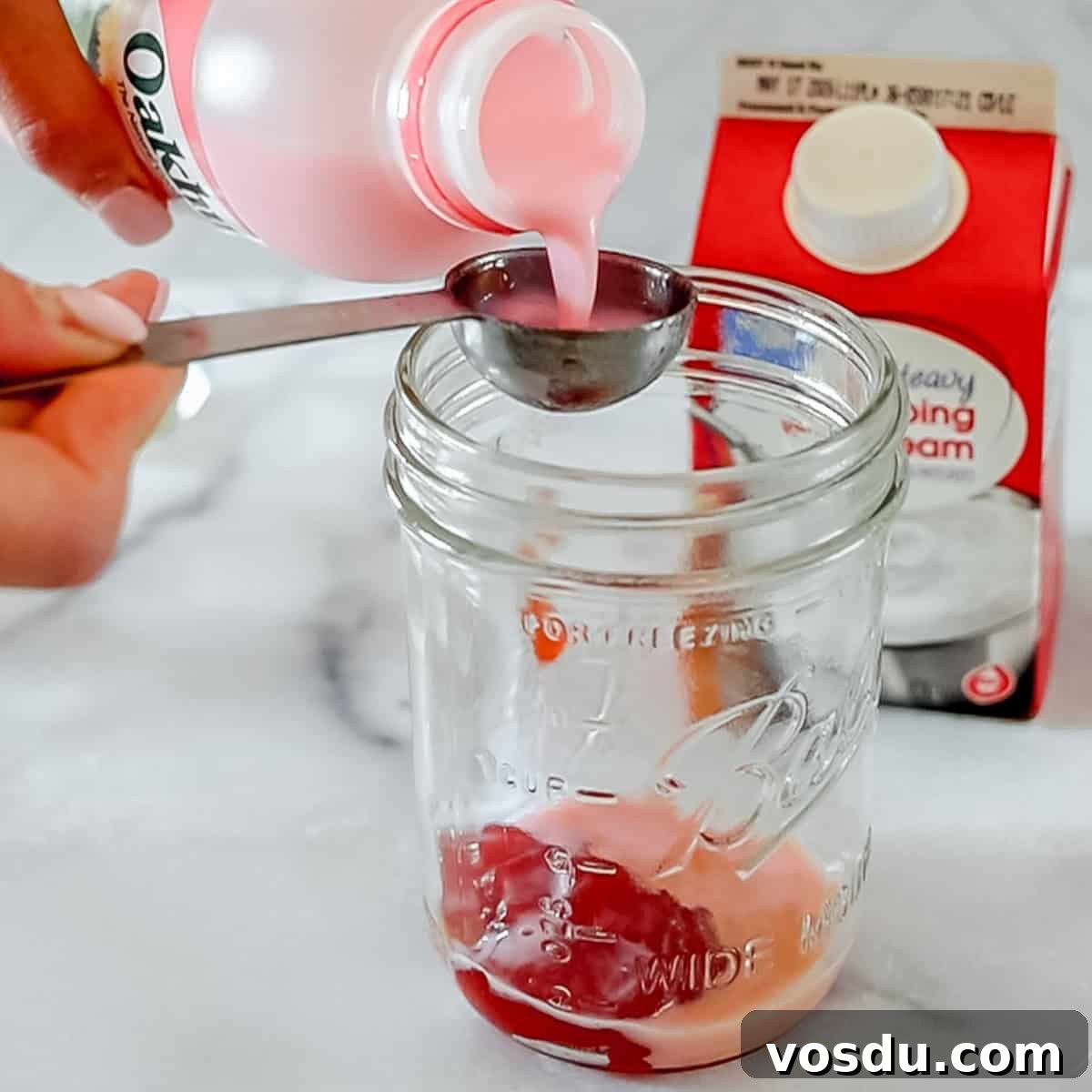 A hand pours pink liquid from a bottle into a measuring spoon over a glass jar, with a carton of heavy whipping cream in the background.