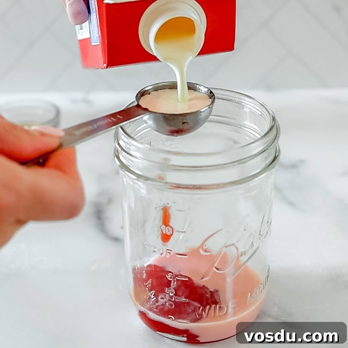 A hand pours cream from a carton into a tablespoon over an empty glass jar, with some cream already inside the jar.