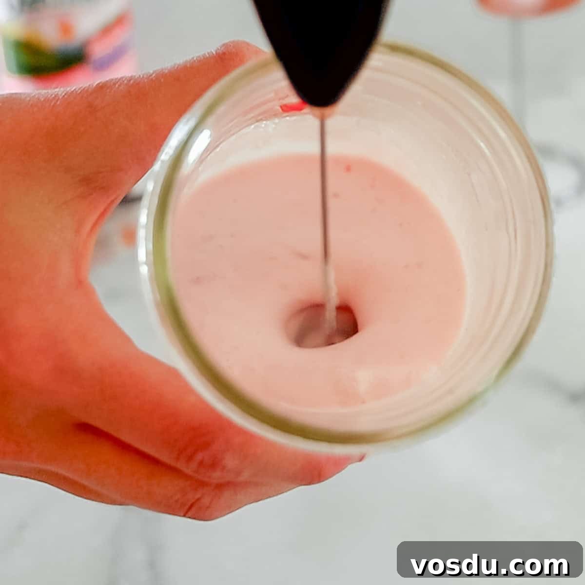 A hand holds a glass filled with pink liquid while a handheld milk frother mixes the contents.